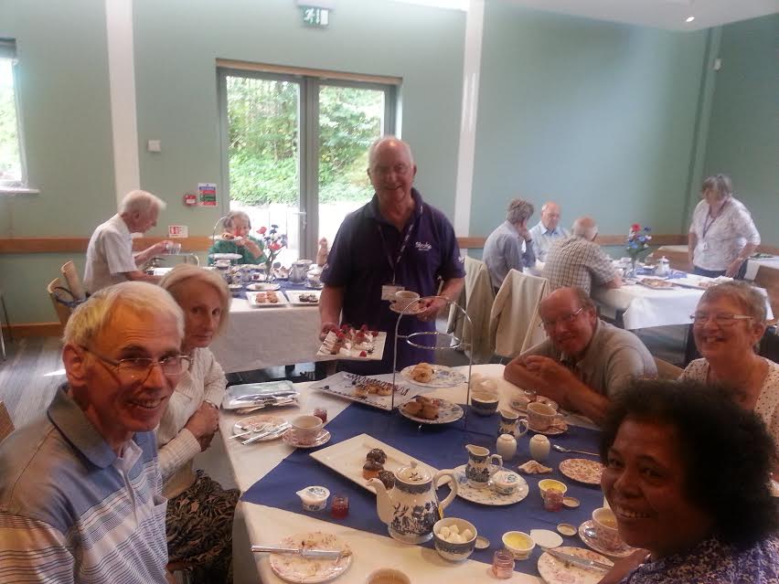 A room full of people sitting at tables, enjoying tea and snacks.