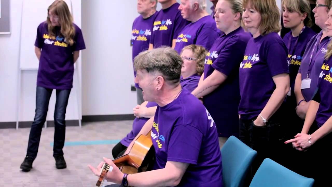 A man playing an acoustic guitar and singing, with several people standing behind him.