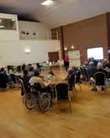 A large hall full of people sitting down on blue chairs and listening to a woman that is talking at the front of the room.