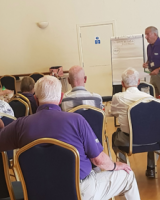 A group of people sitting in a hall, listening to a man standing up in front of them.